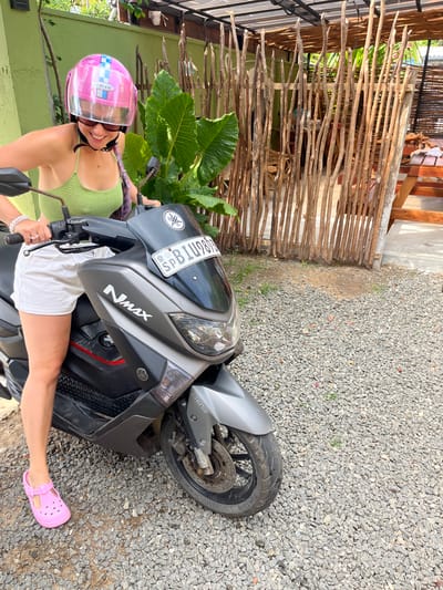 A woman wearing a pink helmet and a green top is sitting on a grey scooter in Kabalana, Sri Lanka. She is smiling and looking towards the camera. The scooter has a license plate with the letters "SPB". The background features a wooden fence and lush green foliage. The ground is covered in small grey pebbles.