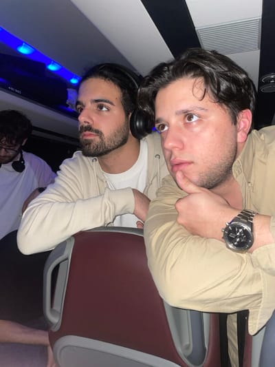 Two young men pose for a photo. The man on top is fair-skinned with brown hair and a watch on his left wrist, wearing a light brown button-down shirt. The man underneath him has a beard and headphones around his neck, wearing a white t-shirt and a light brown zip-up hoodie. Behind them, there are blue lights and part of an airplane seat. The image is rotated, making it appear sideways. Another person wearing headphones is partially visible in the lower center of the image.