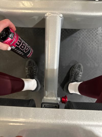 The image shows a person at a gym. The person is wearing maroon leggings, white socks, and black sneakers. They are holding a black and red can of "ABE All Black Everything" energy drink. The can states it has 200mg of caffeine and is cherry slush flavored. The person appears to be sitting on a piece of gym equipment. The flooring of the gym is gray with a checkered pattern. The gym equipment is gray metal. The location is a gym, but the city of Tunbridge Wells, United Kingdom, cannot be determined from the image.