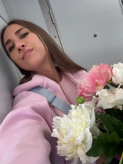 The image features a young woman holding a bouquet of pink and white peonies. She is wearing a light pink hoodie and a light blue strap is visible across her chest. The woman has long, brown hair and is looking at the camera with a slight smile. Her makeup is noticeable, with prominent eyelashes and eyebrows. The background appears to be an indoor setting with a light gray wall that has a metallic hinge and a small, circular fixture on it. The lighting suggests it could be daytime, but there are no specific cues about the time of day or weather. The overall mood is casual and pleasant, with the flowers adding a touch of freshness. The location context is Vyborg, Russia, but there are no specific visual elements in the image that confirm this location. There is no visible text in the image.