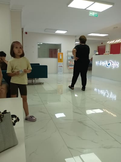 The image depicts the interior of a modern clinic, likely an ophthalmology clinic given the logo "Veni Vidi Göz" which translates to "I came, I saw, Eye" in Turkish. The clinic appears to be located in Ankara, Turkey, as suggested by the Turkish language on the signage. The flooring is polished white marble with a subtle veining pattern, reflecting the bright overhead fluorescent lighting.

In the foreground, to the left, a young girl with dyed hair stands facing forward. She is wearing a light yellow t-shirt and dark brown shorts, and she is holding a few bills in her right hand. Her feet are clad in light purple slip-on shoes with decorative embellishments. Next to her, on the left edge of the frame, a portion of a grey and white patterned handbag is visible.

Further into the background, a woman with dark, curly hair is seen from the back, wearing a black outfit. She is standing with her hands on her hips, looking towards the reception desk. The reception desk itself is white with the illuminated "Veni Vidi Göz" logo prominently displayed. Behind the reception desk, a man is seated, partially visible.

To the left of the girl, there is a row of dark teal seating. Behind the seating, a large window or partition offers a view into another room, where red walls are visible. On the wall to the left of this partition, a red fire alarm pull station is mounted. Above the woman in black, a green exit sign with an arrow pointing left is illuminated on the ceiling. Various informational displays and brochures are visible on a rack to the left of the reception area. The overall impression is one of a clean, well-lit, and professional medical facility.