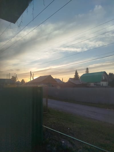 The image depicts a rural street scene in Zavodoukovsk, Russia, at dusk. The sky is a gradient of pale blue and orange hues, with scattered clouds. Several power lines stretch across the frame, intersecting the view of houses and trees. The houses have pitched roofs, with one prominent structure featuring a bright green roof. A dark green fence occupies the foreground, partially obscuring the view of the road and grassy areas. The overall impression is one of a quiet evening in a residential area.