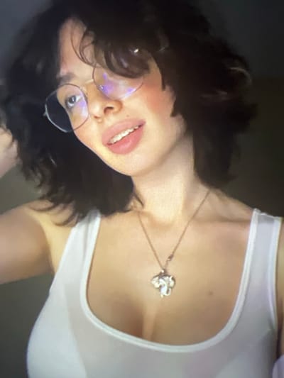 A close-up, low-angle shot shows a young woman wearing glasses and a white tank top. She has dark, wavy hair and is looking at the camera with her mouth slightly open, revealing her teeth. A silver pendant, shaped like an elephant, hangs from a necklace around her neck. The background is dark and out of focus. There is no information about the location in Pyatigorsk, Russia.