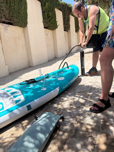 The image shows a man pumping up a stand-up paddleboard (SUP) outdoors. The man, wearing a neon green tank top and navy shorts, is using a black hand pump connected to the valve of the partially inflated blue and white SUP. The SUP is lying on a paved surface, likely a patio or walkway. In the background, there are cream-colored walls and a row of manicured green bushes. The sunlight casts shadows on the ground. A second, deflated paddleboard is visible in the foreground, on the left. A person's legs and feet are visible on the right side of the image, one wearing black sandals. The text "Paddle to Adventure" is visible on the SUP. The scene appears to be in Alicante, Spain, suggested by the style of architecture and vegetation.