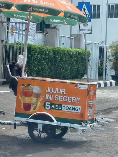 This is a medium shot, outdoor, eye-level, still photograph of a mobile beverage cart selling "Teh Tarik" in South Jakarta, Indonesia. The cart, which is orange and green, is parked on a paved surface, possibly a sidewalk or street. It is equipped with a large umbrella, also orange and green, providing shade.
Prominently displayed on the side of the cart are advertisements for "Teh Tarik KL" (Kuala Lumpur Teh Tarik). The text on the cart reads "JUJUR, INI SEGER!!" which translates to "Honest, this is refreshing!!" and "5 RIBU DOANG!" meaning "Only 5 thousand!". There are also images of two cups of "Teh Tarik" on the cart, one appearing to be a traditional milk tea and the other a lighter colored drink. A QR code and contact information are also visible.
To the left of the cart, a black backpack or bag is hanging on the handle. In the background, there is a dense green hedge and a white building with windows. A pedestrian crossing sign is visible above the umbrella, indicating a designated crossing area. The overall scene suggests a typical street vendor setup in an urban environment.