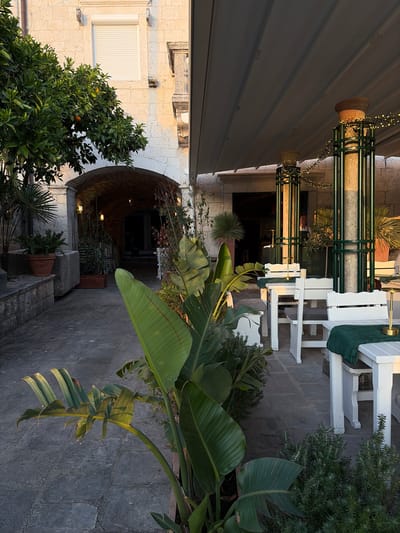 An outdoor seating area of a restaurant in Dobrota, Montenegro, is depicted at dusk. White tables and chairs are arranged under a retractable awning, with green cloths draped over the tables. Decorative string lights adorn the pillars supporting the awning. Lush green plants, including large-leafed tropical varieties, are positioned around the seating area and in pots. An arched entryway leads to an indoor space with soft lighting. A person, presumably staff, is visible standing inside the establishment. The ground is paved with stone tiles, and a stone wall forms part of the building's exterior. An orange tree with visible fruit is situated to the left of the archway.