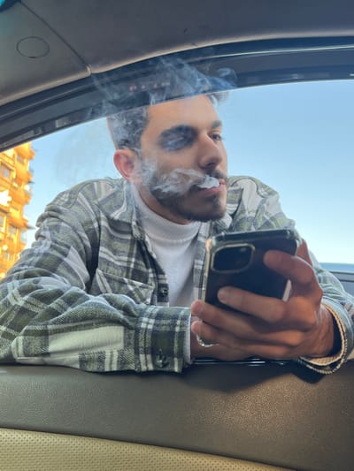 The image features a man captured from a low-angle perspective, looking downwards towards the camera, which appears to be positioned inside a vehicle, possibly pointing upwards through an open sunroof or window. The image itself is rotated approximately 90 degrees clockwise.
The man has dark hair and a beard/stubble. He is wearing a light-colored, possibly white or off-white, turtleneck or crewneck sweater beneath a plaid long-sleeve shirt. The plaid pattern consists of white, light green, and possibly some darker green or gray lines. His mouth is slightly open, and a distinct plume of white smoke is seen actively exhaling from his mouth, swirling around his face and extending to the left side of the frame. His eyes are visible, looking directly towards the camera.
He is holding a dark-colored smartphone, equipped with a clear protective case, in his right hand. The phone is positioned above his head, pointing downwards towards the camera. A ring is visible on his right ring finger.
The immediate setting suggests he is inside a car. Portions of the car's interior, including a light brown or tan upholstered surface (possibly a seat or trim), are visible on the right side of the frame. The edge of a dark-colored sunroof frame or window frame is prominent along the left side of the image.
Beyond the car, a clear blue sky occupies a significant portion of the background. Below what would be the horizon if the image were reoriented, there are blurred architectural structures with orange or brown facades, indicating an urban setting. The overall lighting is bright and natural, suggesting it is daytime. No text is visible in the image.