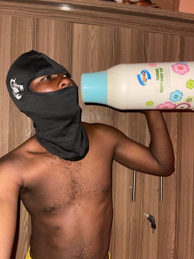 An individual, appearing male and dark-skinned, is positioned indoors within the city of Muchiya, Nigeria. The subject wears a black balaclava featuring a white scorpion logo on the left side, exposing only the eyes, nose, and mouth. The upper torso is bare, displaying a chest with dark hair. A yellow garment is partially visible at the bottom edge of the frame. The individual holds a large, white, opaque plastic bottle with a light blue cap, positioning the cap against their mouth as if consuming its contents. The bottle's label displays colorful floral graphics alongside the text "SMP," "FLOWERS," and "HAPPY EVERY DAY." The background consists of a vertical wooden paneling, against which a set of keys with a black fob hangs from a silver hook on the right side.