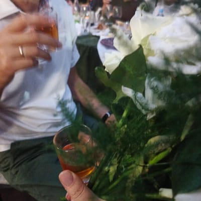 The image shows a close-up of a person's hand holding a small shot glass filled with amber liquid. The person is wearing a white t-shirt and dark pants. A ring is visible on the person's left ring finger. In the background, a bouquet of white flowers is prominently featured. The setting appears to be an outdoor gathering or a restaurant with other people and tables blurred in the background. The location is identified as Svetloye, Russia.
