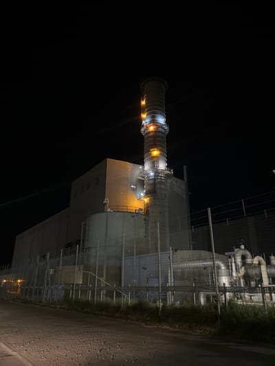 The image depicts a nighttime view of what appears to be an industrial facility in Gorleston-on-Sea, United Kingdom. The main subject is a large structure with cylindrical components and a building-like section illuminated by several lights. The structure is surrounded by fencing, pipes, and vegetation. The ground is dark and textured, possibly asphalt or gravel. The background is primarily black, indicating a night scene. There are no discernible people or specific actions visible.