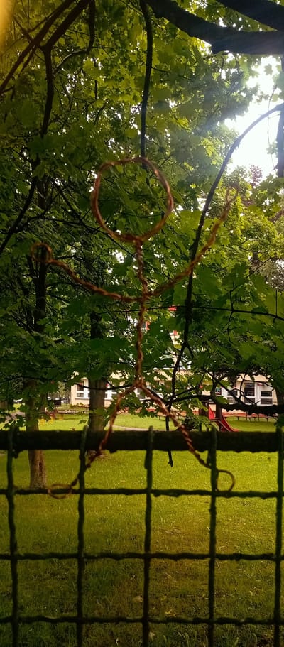 A wire figure resembling a person is suspended from a tree branch, partially obscuring a view of a playground area and a building in Karviná, Czechia. The wire figure's head is a circle, with its body and limbs also made of twisted wire. It is positioned in the foreground, in front of lush green foliage and a grassy area. Behind the figure and foliage, a building with white-framed windows is visible, along with playground equipment including a red slide. A metal fence is in the immediate foreground, partially out of focus, with the wire figure appearing to be in front of or entangled with it. The overall lighting suggests an overcast day.