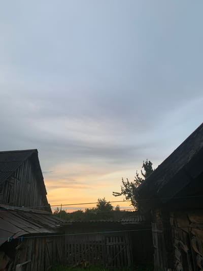 The image captures a rustic scene in Відзаўскі сельскі Савет, Belarus, likely during twilight. Two wooden structures, appearing to be sheds or outbuildings, dominate the foreground, with their weathered wooden walls and corrugated roofs. The structure on the left is partially visible, showing a steep, dark roof. The structure on the right is more prominent, revealing a log-built wall. Between these buildings, a fence made of vertical wooden planks is visible, with a rustic wooden gate. Clotheslines are strung between the structures, with a couple of clothespins attached. Beyond the immediate structures, a line of trees and vegetation stretches across the horizon, silhouetted against a sky with soft, diffused light. The sky itself is a gradient of pale blue and grey, with subtle hints of orange and yellow near the horizon, suggesting either sunrise or sunset. The overall impression is one of rural simplicity and the quiet ambiance of the Belarusian countryside. There are no people or animals visible in the frame.