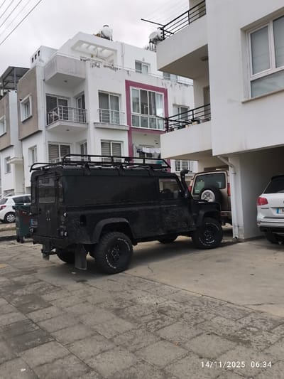 A black off-road vehicle with mud splattered on it is parked on a paved surface in front of buildings. The vehicle has large tires and a roof rack. Two other vehicles are partially visible behind it, along with a green trash can to its left. The buildings have white facades and balconies. The sky is overcast. A date stamp is visible in the bottom right corner.