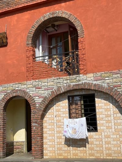 This media file shows an exterior view of a building's facade, focusing on architectural details and features. The building displays a mix of materials, including a rust-colored stucco on the left and a patterned tile facade on the right, composed of cream, yellow, and pink rectangular tiles. Both sections are framed by arched brickwork.

On the left side, a curved opening reveals a window with dark wooden frames and a decorative black metal grate. Inside the opening, there are hints of interior space, including a white pipe. A small, carved wooden object with a metal chain hangs on the stuccoed wall below this opening.

On the right side, a rectangular window is visible behind a black metal grate. A white duvet or blanket, decorated with cartoonish animals in pink and yellow, is hung on a thin wire across the window, suggesting laundry is being dried. The patterned tile wall extends further down, framing another arched opening at the bottom, which shows a glimpse of a yellow-painted interior space.

The lighting suggests it is daytime, with strong sunlight casting shadows, particularly on the lower arched opening. The scene appears to be in a location with a warm climate, possibly Ureki, Georgia, given the context. There are no people visible in the image. The overall impression is one of a residential building with unique architectural design elements.