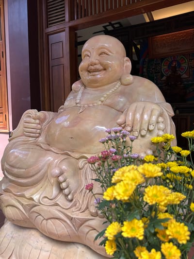 The image shows a statue of a smiling, bald man, likely a depiction of Budai or the Laughing Buddha, a figure associated with good luck and prosperity in Chinese Buddhism. The statue is made of a light-colored, smooth material, possibly stone or ceramic, with a glossy finish. It is adorned with a beaded necklace. The statue is seated, with its hands resting on its lap and its legs crossed. The figure has a jovial expression, with rosy cheeks and a wide smile.
A vibrant bouquet of yellow and pink chrysanthemums is placed in front of and partially obscuring the statue, adding a touch of color and life to the scene. The flowers are in full bloom, with their petals fanned out. The background suggests an indoor setting, possibly a temple or a richly decorated room, with wooden paneling and ornate decorative elements visible. There is a framed piece of artwork with colorful patterns and some text in a language that appears to be Vietnamese on the wall behind the statue. The overall impression is one of tranquility and warmth, evoked by the cheerful demeanor of the statue and the presence of fresh flowers. The lighting suggests it could be daytime, with the ambient light highlighting the statue's features and the flowers. The location context provided, Thôn 5, Vietnam, indicates a Southeast Asian cultural setting.