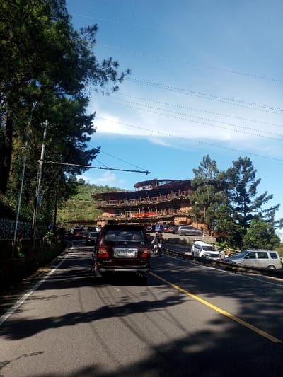 The image depicts a roadside scene on a clear, sunny day in the city of Ka Thaung (Upper), Myanmar. A dark maroon SUV, bearing Indonesian license plate "D 1362 KPJ," is positioned in the foreground, facing away from the viewer, on a two-lane asphalt road. The road features a solid yellow line delineating the right shoulder and a solid white line on the left. Long, irregular shadows from unseen trees or structures are cast across the road surface. A queue of additional vehicles, including various sedans and light-colored cars, extends into the mid-distance behind the prominent SUV, indicating traffic or parked cars. To the immediate right of the maroon SUV, a person on a motorcycle, wearing a light-colored helmet and jacket, is visible.
On the left side of the road, dense green coniferous trees, resembling pines, line an upward slope, partially obscuring the background foliage. A utility pole with multiple overhead wires is visible on the left, extending a horizontal crossarm partially over the roadway.
To the right of the road, a metal guardrail separates the driving lane from an adjacent paved parking area. Several vehicles, including a white SUV and a silver SUV, are parked within this area. Beyond the parking area, a distinctive multi-story building rises on an elevated platform. This structure features a terraced, curved design with at least four visible levels, each incorporating open balconies or verandas secured by railings. The building appears constructed from natural materials, possibly wood and stone. Green trees, similar to those on the left, are interspersed around the building and on the surrounding elevated terrain. The background reveals additional green, hilly landscape under a bright blue sky marked by a few sparse, wispy white clouds, particularly a horizontal streak in the mid-sky.
