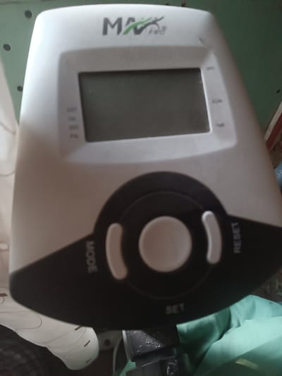 The image shows a close-up of a white and black exercise bike console. The console has a digital display screen at the top, and below it are several buttons, including "MODE", "RESET", and "SET". There is also a large, round central button. The brand name "MAX PRO" is visible in the upper part of the console. The background is blurred and appears to be a room with some green fabric and a white shoe. The image is taken from a slightly elevated angle, looking down at the console. There is no visible indication of the location in Donetsk, Ukraine.