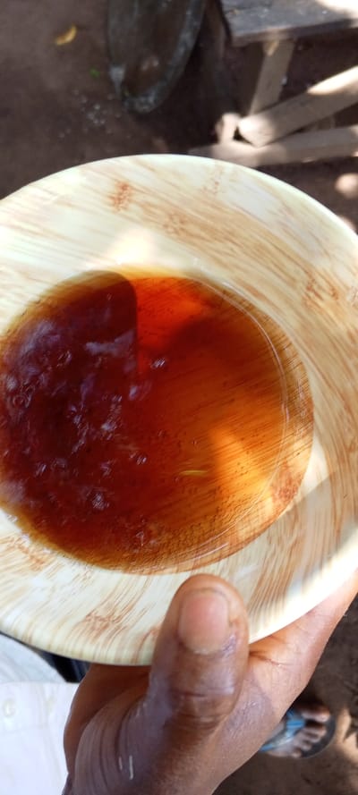 A close-up, top-down perspective depicts a dark-skinned hand holding a light-brown bowl, possibly made of wood or bamboo, exhibiting a textured striped pattern. The bowl contains a translucent dark reddish-brown liquid with visible particulate matter and a single pale yellow, elongate foreign object suspended within it. A thumb from the holding hand rests on the bowl's rim. A portion of a white garment is visible on the subject. The background is out of focus, revealing an uneven ground surface, likely soil, and the distal part of a foot wearing a blue-strapped flip-flop. The scene is illuminated by natural light, indicating an outdoor setting in Akure, Nigeria.