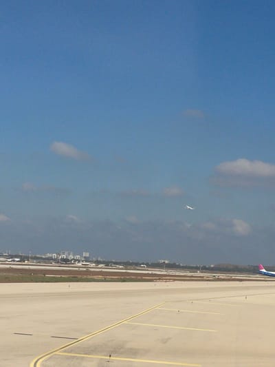 The image captures an aerial view of an airport tarmac in Yehud-Monosson, Israel, under a clear blue sky with scattered clouds. In the foreground, a wide expanse of concrete runway is marked with yellow lines, indicating taxiways or runway edges. The runway stretches towards a distant cityscape visible on the horizon, characterized by low-rise buildings and a few taller structures.
In the middle ground, a commercial airplane is seen ascending, its white fuselage and wings clearly visible against the sky. Further to the right, another airplane, partially visible, appears to be on the runway or taxiing, with its pink tail fin prominently displayed. The environment includes patches of dry vegetation and the infrastructure typical of an airport, such as lights and markers along the tarmac. The overall scene depicts a functional airport environment, likely during daytime.