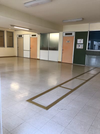 This image captures a brightly lit, empty interior hallway or corridor within what appears to be an institutional building, possibly a school, office, or public facility, located in Agia Paraskevi, Greece. No people are present in the scene.
The floor is covered with light-colored square tiles laid in a diagonal pattern, featuring a polished, reflective surface that mirrors overhead lighting. A prominent detail on the floor is a long, rectangular outline marked by yellow tape or paint, extending from the foreground into the midground. Within this larger rectangle, several smaller yellow rectangular segments are defined, likely serving as designated pathways or social distancing markers.
The walls are predominantly light beige, interspersed with various sections featuring light brown/peach doors, frosted glass panels, and white partitions. On the left side of the hallway, a large, dark green board is mounted. Attached to this board is a white poster providing public health information. The poster clearly displays "CORONAVIRUS" and "COVID-19" in bold text, alongside graphical instructions for hygiene, such as hand washing and mask-wearing, and social distancing guidelines. Some of the accompanying text on the poster is in Greek.
Below the green board, a light brown door is visible with a small sign attached, reading "ΠΡΟΣΩΠΙΚΟ" (Personnel) in Greek, along with a numerical identifier. Further down the hallway, additional doors with frosted glass panels and some with horizontal window blinds are discernible. The ceiling is equipped with linear fluorescent light fixtures, illuminating the space uniformly. The scene conveys a clean, functional, and regulated environment, likely during the daytime.