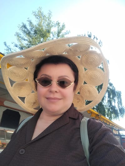 A woman is taking a selfie outdoors, wearing a large, decorative straw hat and sunglasses. She has short, dark hair and is smiling at the camera. She is also wearing a dark brown textured jacket with a button closure and a light green strap is visible over her shoulder, suggesting she is carrying a bag. The background shows trees and a bright, sunny sky, indicating it is daytime. The sunlight is strong, suggesting fair weather. Behind her, there appears to be some sort of outdoor establishment with a yellow awning. The image is cropped to focus on the woman and her accessories. The provided context indicates the location is Zelenogradsk, Russia.
