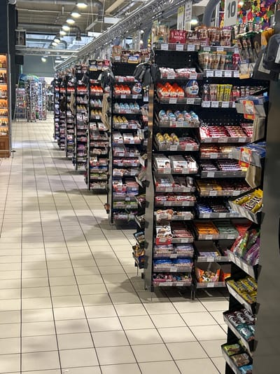 The image depicts an aisle in a supermarket, specifically showing a display of confectionery and beverages. The aisle features multiple shelves stocked with various brands of chocolate bars, candy eggs, coffee products, and other packaged snacks. Notable brands visible include Kinder, Snickers, Nescafe, and various other items that appear to be chocolates and sweets. The shelves are organized in a typical retail manner, with products displayed in their original packaging. The flooring is tiled with a light-colored ceramic tile. Overhead lighting fixtures are visible, illuminating the aisle. In the background, other aisles with more products and shoppers can be seen, suggesting a busy retail environment. The overall impression is that of a well-stocked supermarket confectionery section. The image is dated as being taken in Brovary, Ukraine.