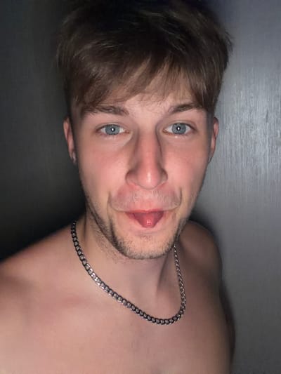 A close-up, chest-up portrait of a young, light-skinned male with short, brown hair and blue eyes is shown. He is wearing a silver chain necklace. His mouth is slightly open with his tongue sticking out. The background is a plain, gray wall with vertical lines. The image appears to be taken in a dimly lit indoor setting. There is no information in the image to suggest its location in Vanino, Russia.