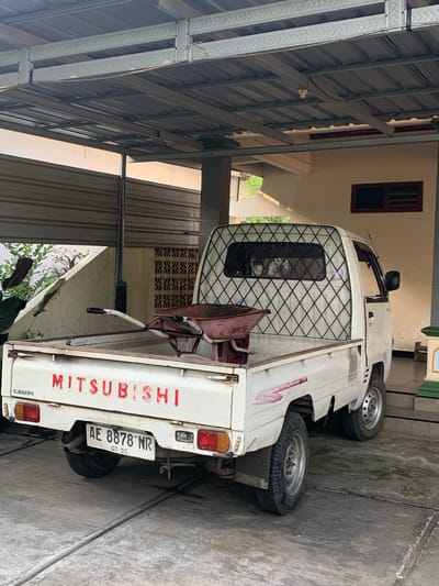 In the city of Tanjungsepreh, Indonesia, a white Mitsubishi pickup truck is parked under a metal-roofed carport. The truck's bed contains a red wheelbarrow. The rear of the truck displays the "MITSUBISHI" logo in red lettering and a license plate with the number "AE 8878 NR". The truck is facing away from the viewer, with its right side visible. The ground is concrete, marked with lines and some wet patches. Behind the truck, a beige wall with a dark-framed window is visible. To the left of the truck, a concrete ramp slopes upwards, and green foliage can be seen. The overall scene appears to be outdoors, in a residential or industrial area, under the shade of the carport.