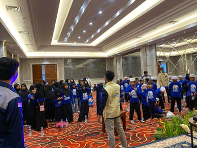 The image depicts an indoor gathering, likely a community event or educational program, taking place in a spacious hall with a red and gold patterned carpet. The ceiling is elaborately decorated with recessed lighting and geometric patterns, suggesting a modern conference or event venue.
In the foreground, a group of children, predominantly girls wearing blue shirts and black hijabs or headscarves, are congregated. Some are interacting with each other, with two girls in the center embracing. Several other children are standing, looking towards the center of the room, while others are seated on the floor. Bottled water is visible on the floor, indicating refreshments were provided.
Adults are also present. A man in a tan vest over a blue shirt, with a logo visible on the vest, appears to be addressing or interacting with the children. Another man, wearing a yellow shirt, stands near a door in the background. Other adults are scattered throughout the scene, some appearing to be supervisors or organizers.
The environment suggests a formal setting, possibly a hotel ballroom or a convention center. The wall in the background features dark patterned panels and large decorative mirrors, reflecting the lighting and the attendees. The presence of multiple participants, the organized arrangement, and the refreshments point towards a planned activity.
The context provided indicates the location is the city of Banjarbaru, Indonesia. While the image itself doesn't offer explicit geographical markers, the overall scene is consistent with such a setting in Indonesia, characterized by the attire of the children and the architectural style of the interior.