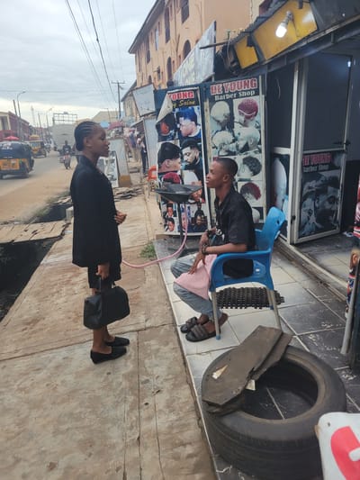 The image depicts a street scene in Egbagu, Nigeria. Two young people are the primary subjects. In the foreground, a person wearing a black outfit and black loafers stands facing away from the camera, holding a black bag. Behind them, another person sits on a blue plastic chair in front of a barber shop. This individual is wearing a black t-shirt, light blue jeans, and dark sandals, and is looking towards the person standing.
The barber shop features posters of hairstyles on its facade. Visible text includes "DE YOUNG Barber Shop". The street appears to be paved with concrete slabs, and there is a discarded tire with some debris inside. The setting suggests an urban or semi-urban environment, with buildings and a street visible in the background, including some three-wheeled vehicles that appear to be auto-rickshaws. The sky is overcast, indicating a potentially cloudy day. The overall mood appears casual and everyday.