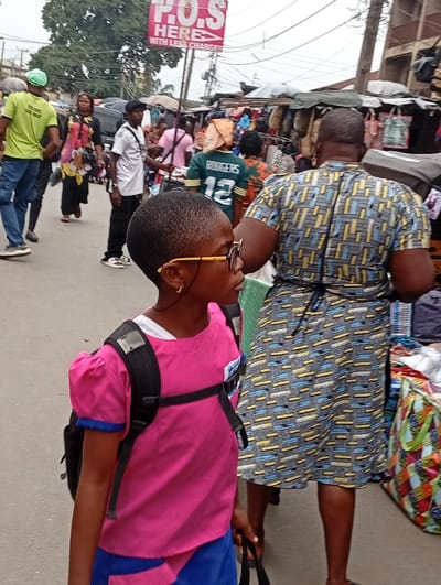The image captures a bustling outdoor market scene, likely during the daytime under an overcast sky, given the diffused light. The setting appears to be a street or pathway lined with vendors.

In the immediate foreground on the left, a young girl is prominently featured, seen in profile looking towards the right. She has short, dark hair and is wearing round, thin-rimmed glasses. Her attire consists of a bright pink short-sleeved top with puffed shoulders and blue trim on the sleeves, paired with a blue skirt or lower garment. She is wearing a black backpack with visible straps over her shoulders and appears to be holding a dark bag in her left hand, which is mostly out of frame.

To the right of the girl, slightly behind her, is the back of an adult woman. She is wearing a distinctive patterned dress with short sleeves, featuring horizontal rectangular shapes in yellow, light blue, and black against a light grey background. A dark tie or string is visible at her waist. She appears to be walking through the market.

The background is filled with numerous other people and market stalls, indicating a busy environment.
On the far left, a man in a light green t-shirt and blue pants, wearing a light green cap, walks away from the camera. Another woman in a dark top with a large floral or patterned design is seen further back.
In the middle ground, several people are visible. One man in a white t-shirt and black cap is walking towards the right, holding what looks like a long pole or stick. Another person is seen from behind wearing a green American football jersey with the name "RODGERS" and the number "12" on the back.
Beyond the people, makeshift market stalls are visible on both sides of the pathway, laden with various goods, including bags, clothing, and other merchandise. The structures supporting these stalls appear somewhat rustic.

Above the crowd, a rectangular red sign with white text is visible. The text reads: "P.O.S HERE WITH LESS CHARGES".

The overall impression is that of a lively and crowded street market, common in many urban areas.