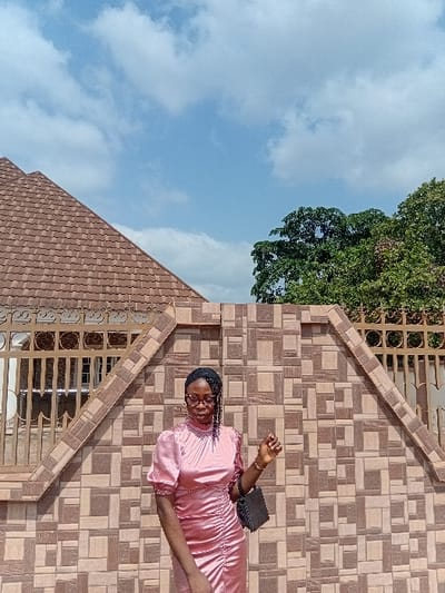 A young woman stands outdoors in Ejule, Nigeria, positioned in front of a patterned tiled wall with brown and beige rectangular and square tiles. She wears a pink, shin-length, short-sleeved, ruched satin dress with a high neckline and puffed sleeves. Her dark hair is braided, and she wears clear-framed eyeglasses. She holds a small black handbag in her left hand, which is bent at the elbow. Her right hand is also bent at the elbow with fingers slightly splayed. Behind the tiled wall, to the left, is a building with a reddish-brown tiled roof. To the right, green tree foliage is visible. The sky overhead is blue with scattered white clouds, indicating daylight conditions.