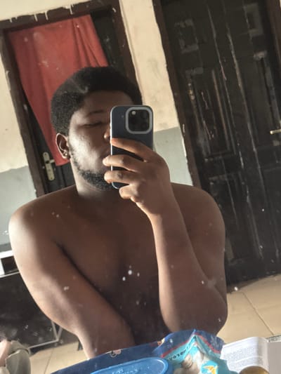 In an indoor setting within the city of Ipetumodu, Nigeria, a male individual with dark skin, short dark hair, and a beard is depicted in a mirror selfie. He appears shirtless, holding a dark-colored smartphone with his right hand, positioned to capture his reflection. A silver stud is visible in his left earlobe. His eyes are partially closed.
The reflected background features a light-colored wall section to the left, partially obscured by a hanging red fabric or curtain. To the right, a dark, paneled wooden door with a gold-colored doorknob is visible. The lower portion of the reflection shows a surface holding a blue plastic container, a blue and white packaged item (resembling wipes or diapers), a folded piece of paper with visible text, and a spherical object. Multiple small, white smudges are present on the mirror's surface, particularly across the reflection of the individual's torso. The environment exhibits signs of wear.