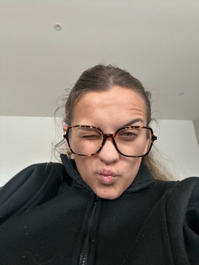 A single female individual is depicted in a close-up, mid-chest to head shot, angled slightly upwards. She has brown hair, pulled back, with visible wrinkles on her forehead. She is wearing large, square-shaped eyeglasses with tortoise-shell frames. Her right eye (viewer's left) is open, looking directly forward, while her left eye (viewer's right) is closed in a wink. Her lips are pursed into a forward, puckered expression. She wears a black, high-collared garment with a visible zipper running down the front. The background is a plain, light-colored ceiling, with a single circular recessed light fixture positioned in the upper center of the frame. The scene is indoors, in Veauchette, France.