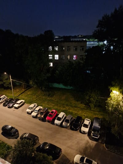 The image depicts a night scene in a residential area, likely in the city of Ерино, Russia, as suggested by the provided context. In the foreground, a row of parked cars is visible on a paved surface, with some cars on the grass verge to the left. The cars are of various models and colors, including black, white, silver, and red. The car furthest to the left appears to be a white station wagon, followed by a black sedan, a dark gray sedan, a white sedan, a silver sedan, a black sedan, a white sedan, a dark gray SUV, a red hatchback, a white sedan, and finally, a dark blue van. In the middle ground, a streetlamp illuminates a patch of grass and a tree. In the background, a multi-story building with illuminated windows is visible amidst dense trees. The overall impression is of a quiet, typical urban night environment. There are no visible people or any ongoing activities captured in the image.