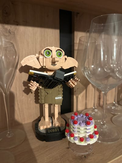 A Lego Dobby figure sits among wine glasses in a wooden drawer. Dobby is brown and beige, holding a black book. A Lego cake also rests on a wine glass. The drawer interior is visible with dark shadows. The wine glasses appear clear and empty. The composition is taken from an overhead angle, showcasing the placement of the Lego figures among the glassware.