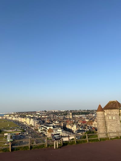 This is a landscape photograph taken in Dieppe, France, from an elevated viewpoint looking down on the town and its coastline. The image is oriented vertically, with the sky occupying the left two-thirds and the town and coastline on the right. The sky is a clear, bright blue with a slight gradient towards a lighter hue near the horizon.
The town of Dieppe stretches along the coast, characterized by a dense collection of buildings with red-tiled roofs, typical of European architecture. Several modern buildings with white facades are interspersed among the older structures. A prominent stone building with a conical roof, possibly a historical landmark or fortification, is visible at the top right of the image, overlooking the town. Below it, a church with a dome is visible.
Along the coast, a street lined with parked cars and some grassy areas is present. There are also several large billboards visible in this area. A hint of the sea or coastline is visible in the distance on the far right, suggesting the town's proximity to the water. The overall scene depicts a coastal town on a clear day, likely in the late afternoon or early evening, given the angle of the sunlight casting light and shadows. There are no specific activities or events clearly depicted, but the presence of cars suggests it is a populated area. The overall emotion conveyed is one of a serene, sunny day in a historic French coastal town. Visible text includes that on the billboards, but it is not legible.