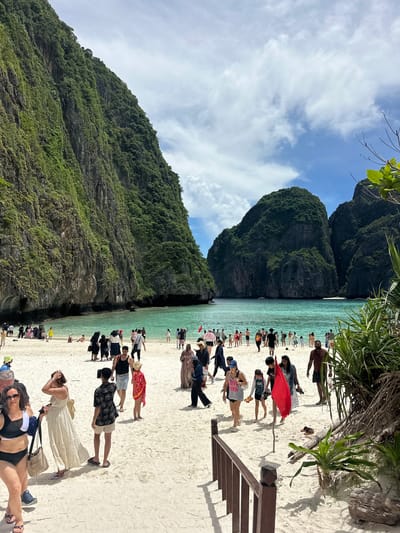 The image depicts Maya Bay, located on Koh Phi Phi Leh island, which is accessible from Ao Nang, Thailand. A crowded white sand beach is visible, with numerous tourists scattered across it. The beach is bordered by turquoise water, and in the background, two large, lush green limestone cliffs rise majestically. The sky above is a bright blue, with scattered white clouds. People are engaged in various activities, including walking, standing, and interacting with each other. Some individuals are wearing swimwear, while others are dressed in casual clothing. The overall scene suggests a popular tourist destination on a sunny day.