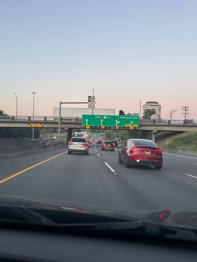 The image shows a highway scene in Burnaby, Canada, during dusk. The perspective is from inside a vehicle, looking forward. Traffic is moderate, with several cars on the multi-lane highway. The sky is a pale pink and blue, indicating the transition from day to night.

A large green overhead highway sign indicates exits for "Hope" (East) and "Willingdon Ave" (Exit 29). The "Willingdon Ave" exit is marked as "ONLY". Smaller yellow signs below the main sign show height restrictions, one indicating "4.3 m" and another with a graphic of a truck and a car, also likely indicating height or width restrictions.

Several vehicles are visible. In the foreground, a dark-colored vehicle's dashboard and windshield are prominent. Slightly ahead and to the right is a red Tesla Model X. To the left of the Tesla and further ahead is a silver SUV. Other cars are visible in the middle and far distance, some appearing to be part of a traffic jam.

An elevated overpass spans the highway in the middle ground. Underneath the overpass, more highway infrastructure and trees are visible. In the background, a tall, distinctive building, possibly an office tower or control tower, stands against the sky, along with power lines. The highway has yellow and white lane markings. The scene is consistent with a freeway environment in a metropolitan area.