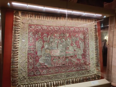 This image captures a close-up view of an intricately embroidered tapestry displayed within a glass case. The tapestry, predominantly in shades of red and muted green, features a central scene depicting a religious gathering with multiple figures, possibly a biblical or historical narrative. The figures are rendered in a stylized, somewhat flat manner, typical of ancient textile art. Angels with wings are visible, as are robed individuals surrounding a central element that could be a bed or an altar. Decorative borders frame the central imagery, incorporating floral and geometric motifs. The tapestry is further embellished with rows of tassels along its bottom and side edges, suggesting it may have been part of a larger garment or used as a ceremonial banner. The display case is illuminated by overhead lights, creating reflections on the glass surface. The background shows a dimly lit museum environment, with hints of exhibit walls and architectural elements, including a column and the orange-hued wall of the exhibition space. Although the image focuses on the tapestry, the context suggests it is an artifact housed in a museum. The prompt mentions Kalambaka, Greece, which is known for its Meteora monasteries, often housing significant religious artifacts. Given this, the tapestry likely originates from or is displayed in a cultural institution within or near Kalambaka, Greece, though the image itself does not contain explicit geographical markers.