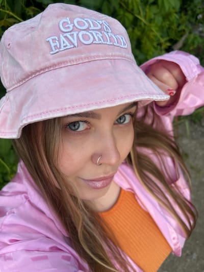 A young woman with light brown hair and blue eyes is seen from the chest up, wearing a pink bucket hat and a shiny pink jacket over an orange top. The hat has embroidered white lettering that reads "GOD'S FAVORITE". She is wearing a silver nose ring. Her left hand is visible, with red nail polish on her fingernails. The background consists of green foliage and a blurred surface that appears to be concrete. The lighting suggests it is daytime. The scene is likely outdoors, possibly in a park or garden setting. The location in Novopokrovskaya, Russia cannot be determined from the visual information provided.