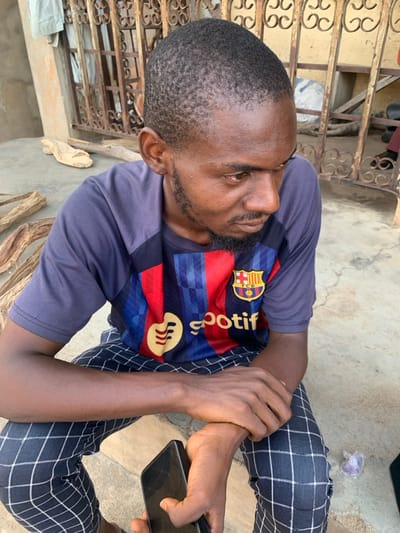 A young adult male, appearing to be in his late twenties to early thirties, with dark skin, short dark hair, and a short beard and mustache, is seated on a light-colored concrete surface. He is wearing a short-sleeved dark blue and striped red/blue sports jersey featuring the Spotify logo and FC Barcelona crest, along with dark-colored trousers displaying a white grid pattern. His gaze is directed downwards and to his right. His left hand holds a black smartphone, while his right hand rests on his left forearm. Behind him, a light-colored building wall is visible, along with a dark brown or black ornate metal gate or grille. Several pieces of dried, light-colored wood are scattered on the ground to his left, and a small crumpled piece of plastic or paper is on the ground to his right. The scene is located in Okene, Nigeria.