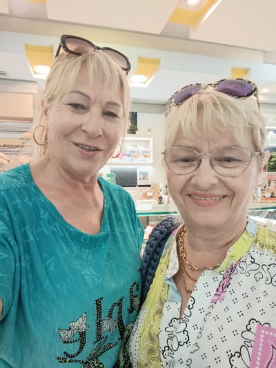 Two women are posing for a selfie, with the woman on the left in the foreground and the woman on the right slightly behind her and to the right. Both women appear to be in their 50s or 60s, with blonde hair. The woman on the left is wearing a turquoise t-shirt with a pattern of letters and a design of leaves and what looks like a rabbit. She has large hoop earrings and sunglasses perched on her head. The woman on the right is wearing glasses and a colorful patterned shirt with yellow, white, and blue sections, featuring a paisley print and small black dots. She is wearing a gold chain necklace. Both women are smiling, suggesting a positive and friendly mood.
The setting appears to be indoors, possibly a store or market, given the shelves with products visible in the background. There are bright lights overhead, possibly indicating daytime. The overall impression is one of a casual outing between friends or family members. No specific time of day or weather conditions are discernible. There is no visible text in the image. The location context of Odivelas, Portugal, does not provide any specific visual cues within the image itself.