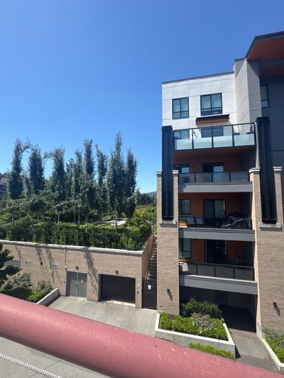 The image shows a modern apartment building with multiple balconies, situated in Port Moody, Canada. The building features a combination of light brick and grey and white paneling, with orange accents visible on some balconies. The balconies have glass railings and black vertical supports. Below the balconies, there is a building with grey double doors and a large rolling garage door. Lush greenery, including numerous tall, slender trees and various shrubs, covers the area behind a black metal fence, suggesting a well-maintained landscape. The sky is clear and blue, indicating a sunny day. In the foreground, a red railing and some rope are visible, suggesting the image was taken from an elevated viewpoint. The overall scene depicts a residential area with contemporary architecture and abundant plant life.