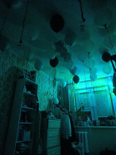 The image depicts an indoor scene, possibly a bedroom, decorated with numerous balloons suspended from the ceiling. The room is illuminated with blue and green lights, creating a moody and somewhat surreal atmosphere. There is a patterned wallpaper on the wall, and a shelving unit is visible on the left side of the frame, filled with various items. A dresser with drawers is located in the center, and a window with curtains is present on the right. The overall impression is that of a decorated space, possibly for a celebration. There is no indication of the city of Kaliningrad, Russia in the image.