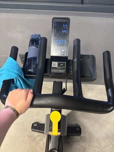The image shows a close-up view of a stationary exercise bike. The digital display reads "77" and "44" at the top, and "0653" below. A water bottle in a holder is visible on the left side of the bike's handlebars. A light blue towel is draped over the lower part of the handlebars. A person's hand, adorned with a bracelet, is holding onto the handlebars. The background consists of neutral-colored walls. The image does not depict any location in the city of Córdoba, Spain.