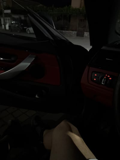 The image shows the interior of a car at night. The focus is on the dashboard and door panel, which have red accents. A person's leg and arm are visible on the right side of the frame, suggesting they are sitting in the driver's seat. The dashboard has several buttons illuminated with red light, including what appears to be a central locking button and a heated seat button. The time of day is clearly night, and the setting appears to be a residential area, possibly Zverino, Bulgaria, given the location context. The overall mood is somewhat dark and intimate, typical of a nighttime car interior. There is no visible text in the image.