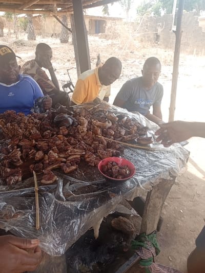 The image shows a group of four men gathered around a table piled high with a large quantity of grilled meat. The men are seated in a partially covered outdoor setting, with a thatched roof overhead, constructed from a wooden frame and supported by wooden poles. The table is covered in a plastic sheet, on which sits an abundance of grilled meat, likely pork. The meat is arranged on the table in a haphazard manner, with some pieces neatly stacked and others spread out in a more casual fashion. A red bowl filled with additional pieces of meat rests on the table. The men are dressed in various casual attire. Two of the men are wearing shirts with logos on them, while one man is wearing a white shirt and another man is wearing a blue shirt. The men appear to be enjoying the meal together, as evidenced by the fact that they are all looking at the food and engaging in conversation with each other. The environment is relaxed and convivial, with the men seeming to be enjoying the meal and each other's company. The location is Assai Sabongida, Nigeria, based on the latitude 8.864 and longitude 8.442.