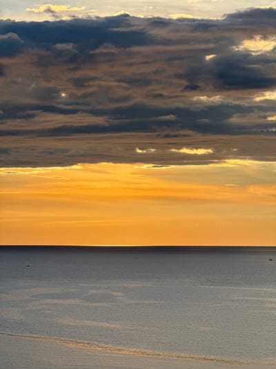 This image captures a serene sunset over the sea, likely near the city of Lý Sơn Special Administrative Region, Vietnam. The sky is a dramatic display of color, transitioning from a pale yellow at the horizon to vibrant orange and then to dark, textured clouds in the upper portion of the frame. The sun, low on the horizon, casts a bright, narrow reflection across the calm, deep blue sea. The water is largely featureless, with subtle variations in light and shadow suggesting gentle ripples. A few distant, small boats are visible as dark silhouettes on the vast expanse of water, contributing to the sense of scale and tranquility. The overall scene evokes a peaceful and majestic natural spectacle.