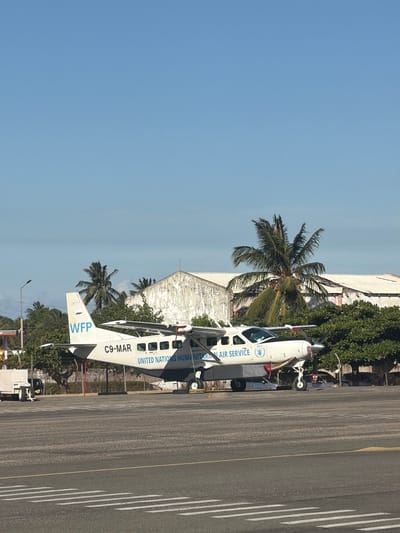This vertically oriented image captures a white propeller aircraft parked on an asphalt tarmac at an airport, identified as being in Pemba, Mozambique. The aircraft is a small, twin-engine model with a clear registration number "C9-MAR" on its fuselage. Prominently displayed on its tail and side are the blue "WFP" (World Food Programme) logo and the text "UNITED NATIONS HUMANITARIAN AIR SERVICE," indicating its role in humanitarian aid.

The plane is situated next to a section of tarmac with distinct white parallel lines and a yellow line, characteristic of airport runway or taxiway markings. To the left of the aircraft, a line of tall palm trees and dense green vegetation thrives, suggesting a tropical environment. Behind these trees and parts of the aircraft, a light-colored, somewhat weathered building, likely an airport hangar or facility, is visible.

The sky is a clear, light blue, indicating bright, sunny weather during the day. Strong sunlight creates visible shadows on the tarmac. No individuals are clearly depicted engaging in specific activities, but some small ground support equipment, including what appears to be passenger steps, is partially visible near the aircraft. The scene portrays an operational humanitarian aircraft in a tropical airport setting.